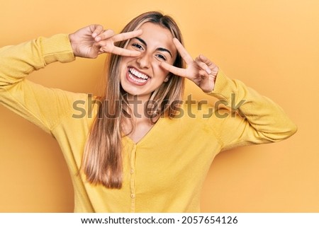 Beautiful hispanic woman wearing casual yellow sweater doing peace symbol with fingers over face, smiling cheerful showing victory 