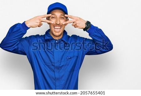 Bald man with beard wearing builder jumpsuit uniform doing peace symbol with fingers over face, smiling cheerful showing victory 