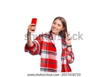 Lovely smiling woman in red is taking a selfie while showing v gesture over white background.