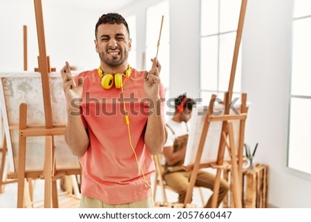 Young hispanic man at art studio gesturing finger crossed smiling with hope and eyes closed. luck and superstitious concept. 