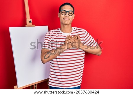 Young african american guy standing by painter easel stand smiling with hands on chest, eyes closed with grateful gesture on face. health concept. 