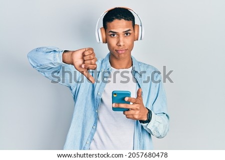 Young african american guy using smartphone wearing headphones with angry face, negative sign showing dislike with thumbs down, rejection concept 