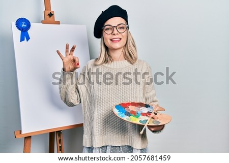 Beautiful caucasian woman drawing with palette on easel stand winner first place doing ok sign with fingers, smiling friendly gesturing excellent symbol 