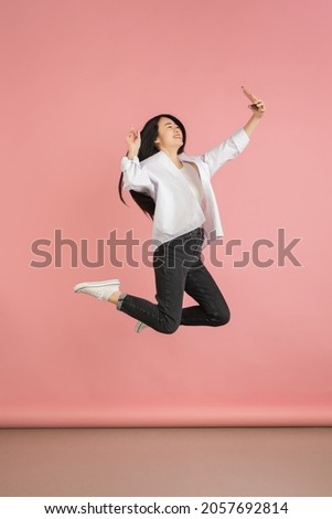 Jumping high, taking selfie. Asian young woman's portrait on pink studio background. Beautiful female model in casual style. Concept of human emotions, facial expression, youth, sales, ad.