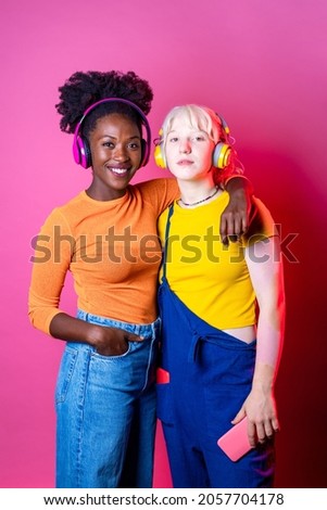 Two young diverse multicultural women best friends posing isolated background having fun laughing holding smartphone wearing wireless headphones