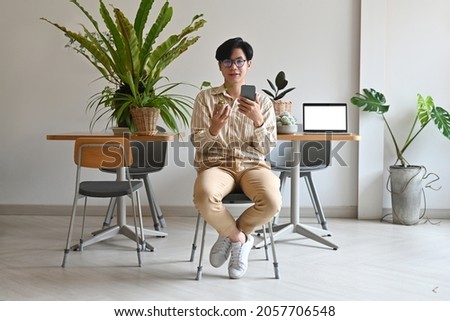 Photo of a young man showing a small plant and smartphone in hand while sitting over the working desk surrounded by a computer laptop and potted plant as a background.