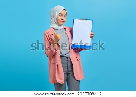 portrait of cheerful beautiful asian woman holding clipboard showing thumbs up on blue background