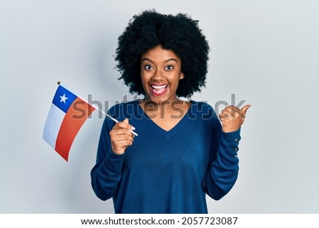Young african american woman holding chile flag pointing thumb up to the side smiling happy with open mouth 