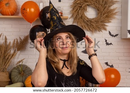 A beautiful woman in a black dress puts on a witch hat on a background decorated for Halloween. Preparing to celebrate the Feast of All Saints and make Jack's lantern.