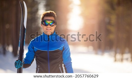 Portrait happy male athlete with cross country skis in hands and goggles, training in snowy forest. Healthy winter banner.