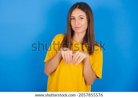 Young european brunette woman wearing yellow T-shirt on blue background makes bunny paws and looks with innocent expression plays with her little kid