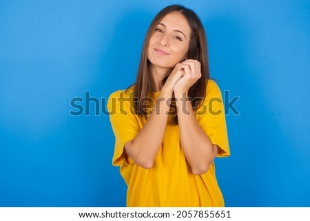 Charming serious Young european brunette woman wearing yellow T-shirt on blue background keeps hands near face smiles tenderly at camera
