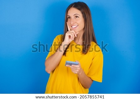 Image of a thinking dreaming Young european brunette woman wearing yellow T-shirt on blue background using mobile phone and holding hand on face. Taking decisions and social media concept.