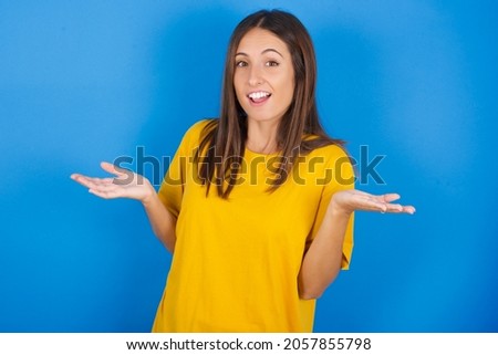 So what? Portrait of arrogant Young european brunette woman wearing yellow T-shirt on blue background shrugging hands sideways smiling gasping indifferent, telling something obvious. Royalty-Free Stock Photo #2057855798