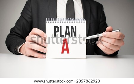 man writes in a notebook with a silver pen and hand holding card with text PLAN a. grey background, front view. business and education concept.