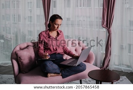 a Caucasian female psychologist in a pink shirt is sitting on the couch and working on a laptop