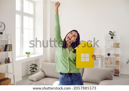 Happy single young woman who finally has enough money to buy new home or pay off mortgage standing in living room, holding symbolic paper house, raising hand up and shouting YES. Real estate concept Royalty-Free Stock Photo #2057885573