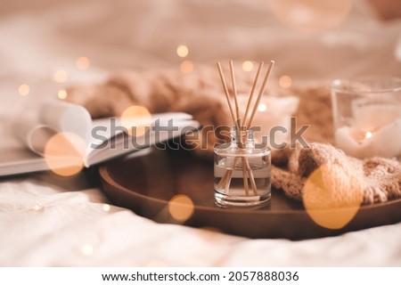 Home perfume in glass bottle with wood sticks, scented burn candles, open paper book and knit wool textile on ray in bedroom close up. Aromatherapy cozy atmosphere lifestyle. Winter warm xmas season.  Royalty-Free Stock Photo #2057888036