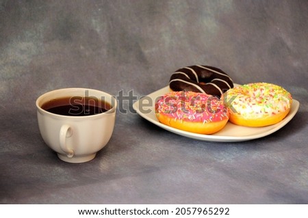 A cup of black tea and a plate with three glazed donuts on a gray background. Close-up.