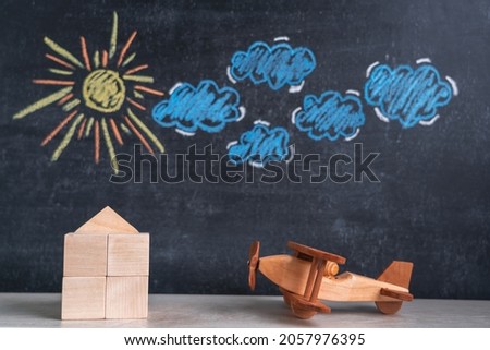 A wooden house and a toy airplane stand against a chalk board with a picture of the sun and clouds. The concept of returning home from a trip
