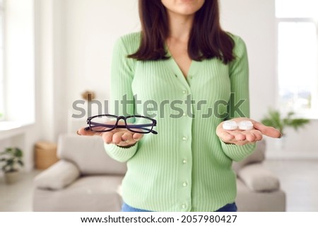 Woman making choice between glasses and eye lenses. Young girl who's holding eyeglasses and container box with pack of modern color lenses is trying to choose which to wear. Cropped shot, close up Royalty-Free Stock Photo #2057980427