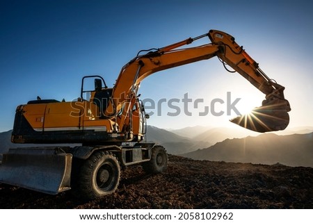 Excavator is working on the hill at sunset. Excavators are heavy construction equipment consisting of a boom, dipper (or stick), bucket and cab on a rotating platform.