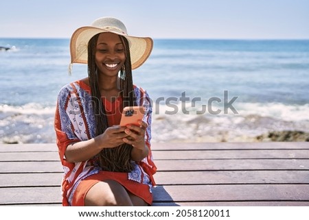 Young african american tourist woman using smartphone sitting on the bench at the beach. Royalty-Free Stock Photo #2058120011