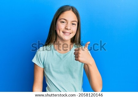Beautiful brunette little girl wearing casual white t shirt doing happy thumbs up gesture with hand. approving expression looking at the camera showing success. 