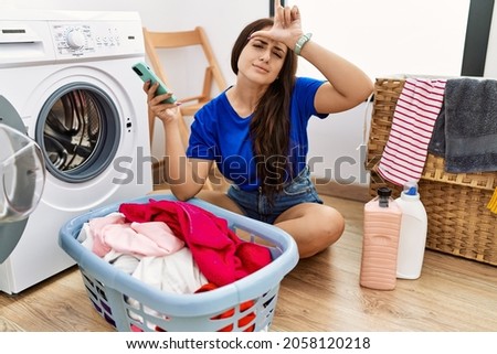 Young brunette woman doing laundry using smartphone making fun of people with fingers on forehead doing loser gesture mocking and insulting. 