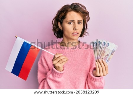 Young brunette woman holding russia flag and rubles banknotes clueless and confused expression. doubt concept. 