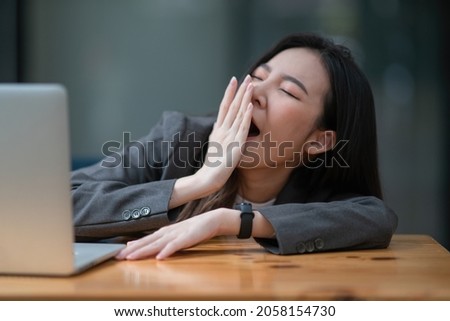 Young businesswoman yawning at a modern office desk in front of a laptop, covering her mouth out of courtesy
