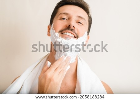 Close-up portrait of handsome positive man with towel on shoulders applying moisturizing shaving foam, smiling guy looking at camera and doing his daily shave, skin care, male beauty. Royalty-Free Stock Photo #2058168284