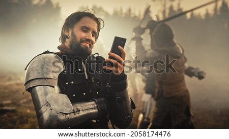 Portrait of Handsome Medieval Knight Using Smartphone on Battlefield, Smiling. Funny Concept: Armored Warrior Having Fun, Ordering Online, Betting, Investing, doing E-commerce. War is Raging Royalty-Free Stock Photo #2058174476
