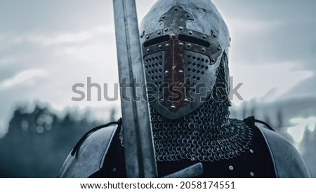 Medieval Knight Wearing Armour and Helmet, Draws Sword from Shearh, Ready to Fight, Kill His Enemy in Battle. Warrior Soldier on Battlefield. War, Invasion, Crusade. Cinematic Historic Reenactment Royalty-Free Stock Photo #2058174551