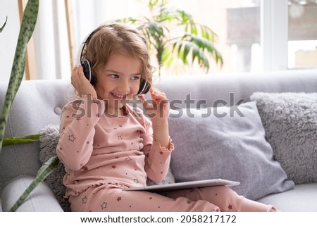 Smiling happy little kid caucasian girl using tablet in wireless headphones in couch at home.