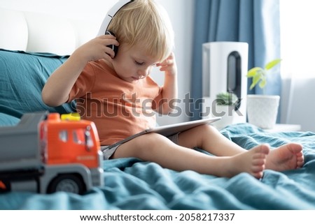 Smiling happy little kid boy using tablet in wireless headphones in bed at home.