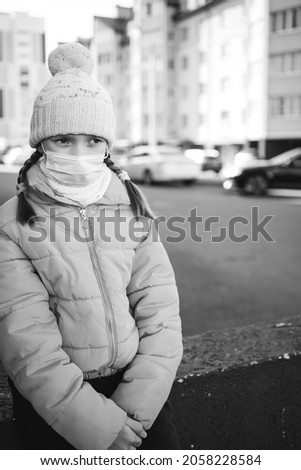 sad girl in a yellow jacket and medical mask sits on a concrete slab on the background of the autumn city. black and white photo