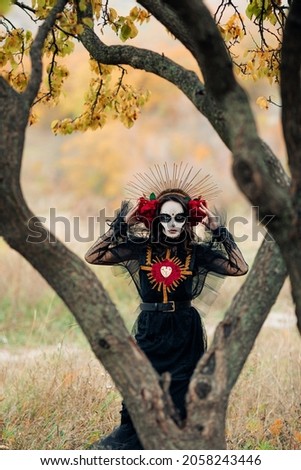 Young woman with sugar skull makeup and red roses dressed in black costume of death as Santa Muerte is against background of autumn forest. Day of the Dead or Halloween concept.