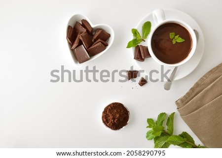 Hot chocolate in white ceramic mug, pieces in heart-shaped bowl and chocolate powder in bowl on white table. Top view. Horizontal composition.