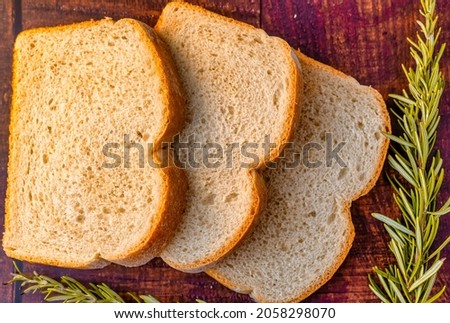 Drei Scheiben frisch geschnittenes Brot auf rustikalem braunem Hintergrund mit einer Rosmarinpflanze in der Dekoration. Royalty-Free Stock Photo #2058298070