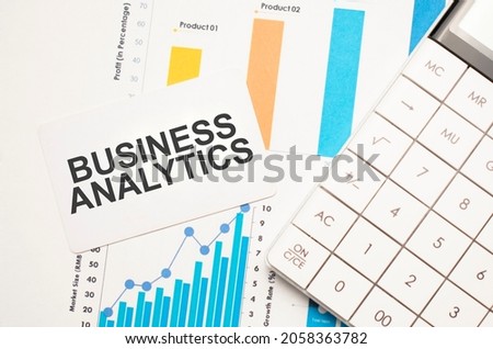 business analytics, text concept. Office workplace table with calculator, graphs, reports and the text business analytics on a small piece of paper on multicolored background.