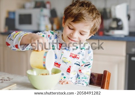 Happy little blond kid boy eating cereals for breakfast or lunch. Healthy eating for children in the morning. Child in colorful pajama nightwear having breakfast with milk and oat granola muesli Royalty-Free Stock Photo #2058491282