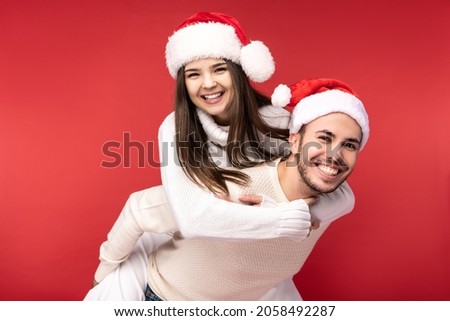 Photo of sweet couple in Christmas hats, happy about holidays, she jumped on his back. Male and female are in love, look blessed and smile, isolated over red background