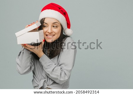 Photo of cute smiling female in Christmas hat is curiously opening present. Wears grey shirt, isolated grey color background