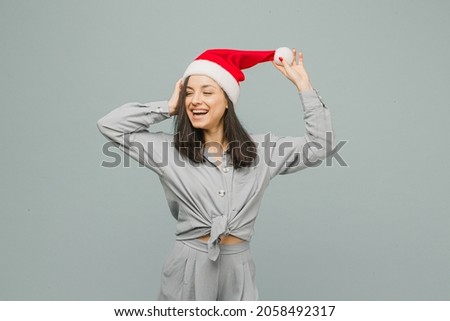 Photo of cute smiling female in Christmas hat looks happy. Wears grey shirt, isolated grey color background