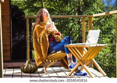 Pretty white girl is sitting relaxing on balcony wrapped in yellow plaid holding cup of tea coffee. Young brunette woman works remotely on laptop, shopping online. Sweet home in a wood at autumn day. Royalty-Free Stock Photo #2058545816
