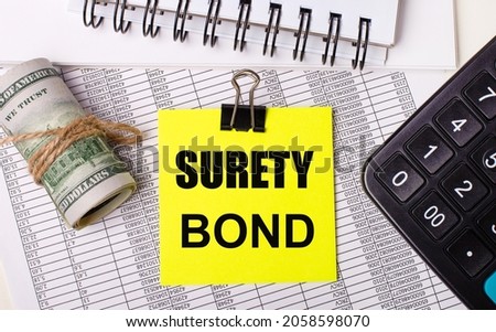 On the desktop there are reports, notepads, a calculator, a cash and a yellow sticker with the text SURETY BOND. Business concept Royalty-Free Stock Photo #2058598070