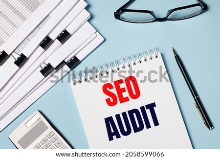 On a light blue background there are documents, glasses, a calculator, a pen and a notebook with the text SEO AUDIT. Close-up of the workplace. Business concept