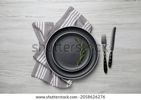 New dark plates with rosemary and cutlery on light wooden table, flat lay