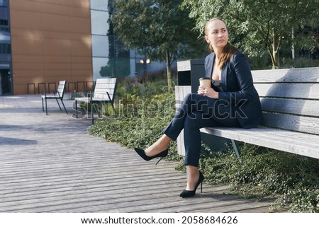 Pensive european business woman has coffee break, sits outside of office on bench with takeaway cup in hand, looks aside, wears business suit, high heels and deep neckline blouse, has ponytail